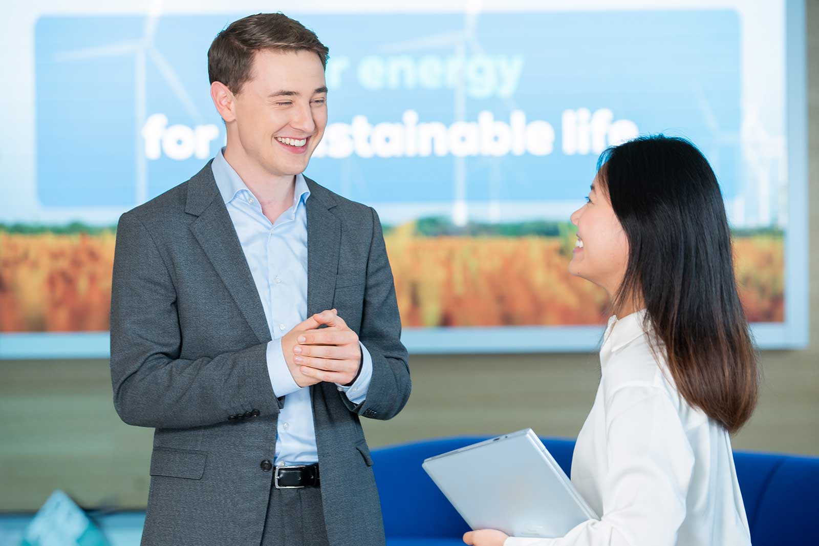 A man in a suit converses with a woman holding a tablet in a modern office with a sustainable energy backdrop.