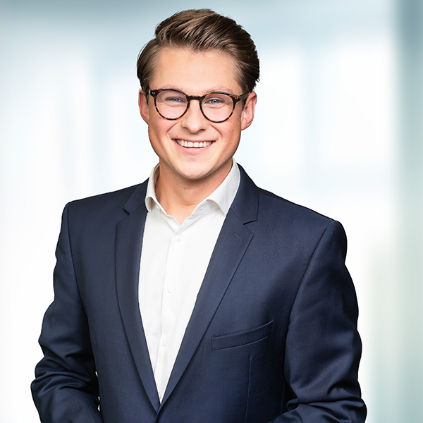A young man in a dark blue suit and white shirt stands smiling against a bright, blurred background.
