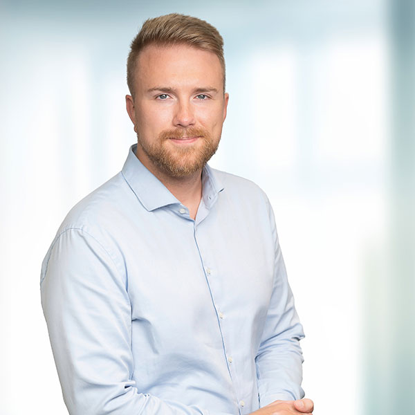 A man in a light blue shirt stands against a blurred background, with arms crossed and a neutral expression.