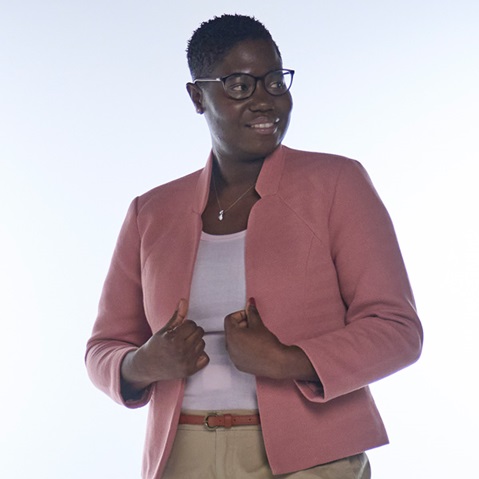 A person wears a light pink jacket over a white top. They pose with both hands on their waist.