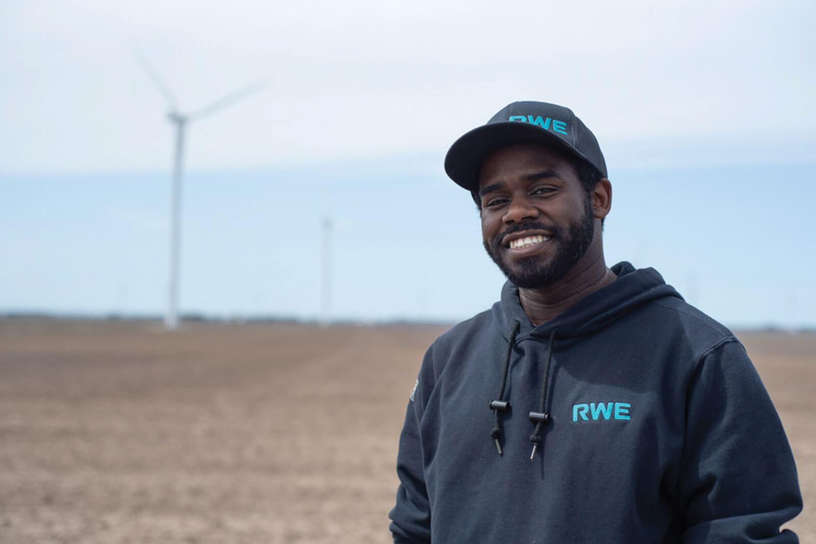 A man in an RWE hoodie stands in a barren field with wind turbines in the background.