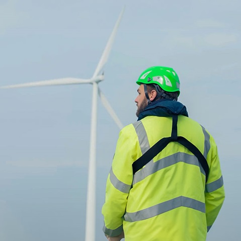 A man in a green safety helmet and reflective jacket stands in front of a wind turbine.