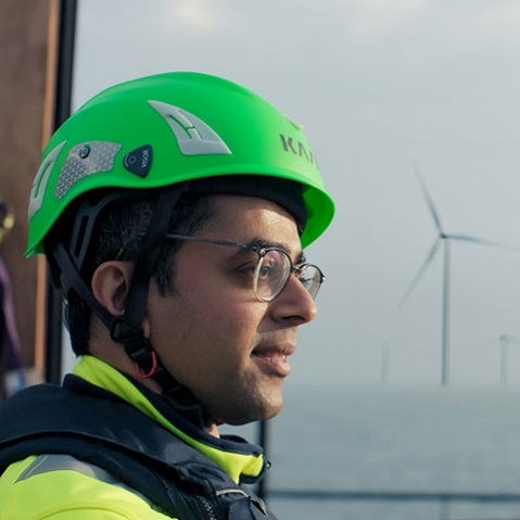 A man in a bright green helmet stands by the water, with wind turbines visible in the background.