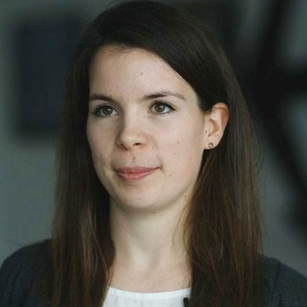 Eine Person mit langen, dunklen Haaren trägt ein graues Oberteil und Ohrringe. Der Hintergrund ist unscharf.