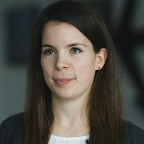 A person with long dark hair is wearing a grey top and earrings. The background is blurred.