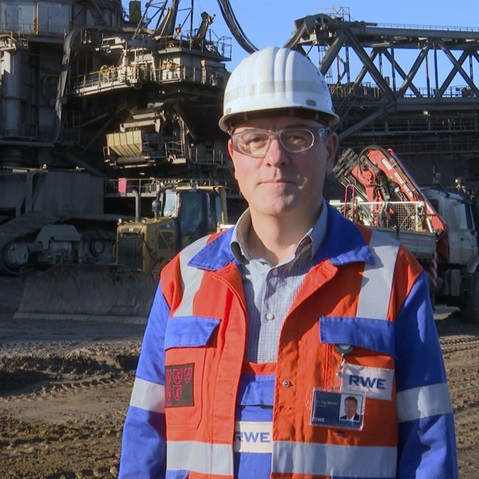 A worker in an orange-blue safety suit and helmet stands before heavy machinery and vehicles on an industrial site.