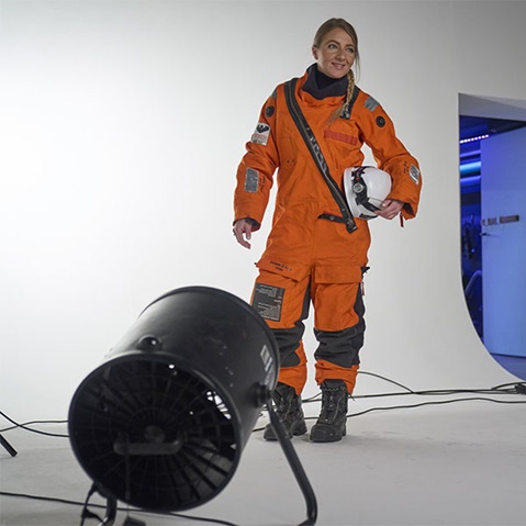 A person in an orange space suit is holding a helmet. A fan is placed in the foreground.