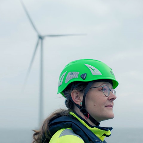 A person wearing a green helmet and a yellow jacket stands in front of a wind turbine on a cloudy day.