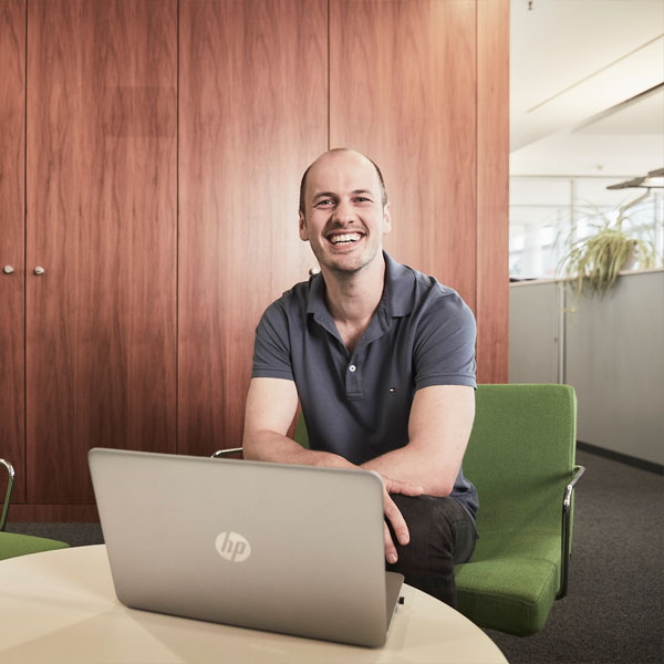 A man sits in a modern office on a green chair, working on a laptop on a round table.