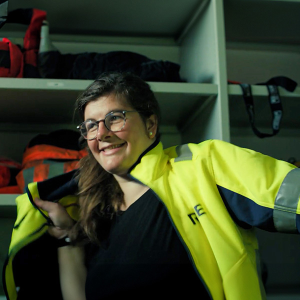 A person is putting on a bright yellow jacket, with a shelf of backpacks visible in the background.
