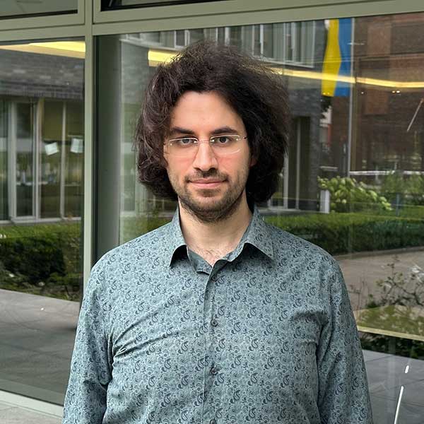 A man with curly hair is wearing a patterned shirt and stands in front of large windows in a modern building, portrait of Dr Farshid Balaneji.