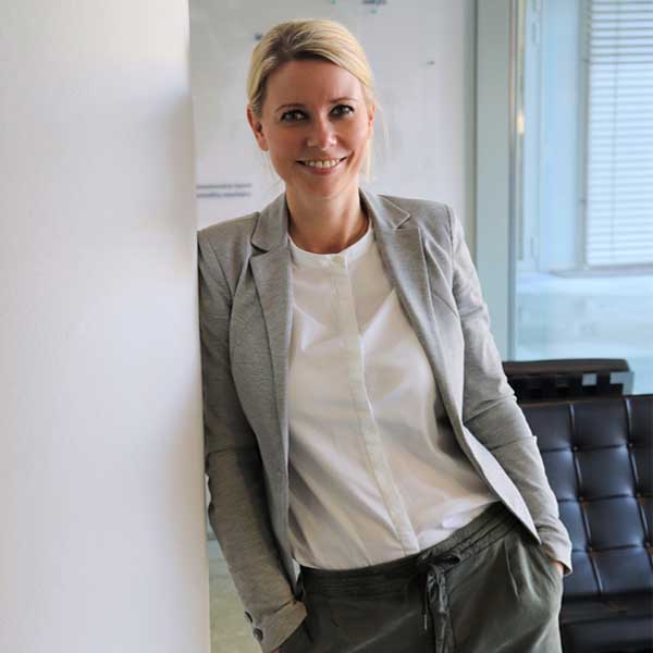 A person in a grey blazer and white blouse leans against a wall in a modern office, portrait of Madlen Krembel.