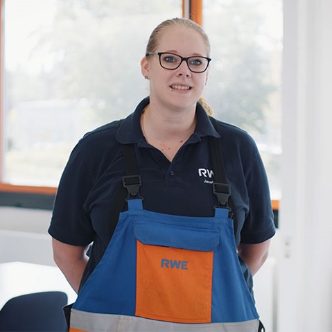 Eine Person steht mit den Händen hinter dem Rücken in Arbeitskleidung und trägt eine blau-orange RWE-Latzhose sowie ein dunkelblaues Poloshirt vor einem Fenster.