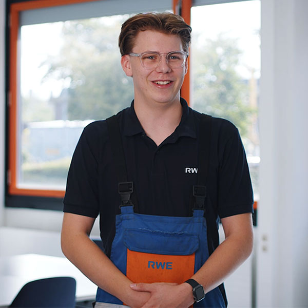 Ein Arbeiter in schwarzem Polo mit RW und blau-orangefarbener Latzhose mit RWE steht drinnen an einem Fenster mit orangefarbener Umrandung, die Hände gefaltet und mit Smartwatch.