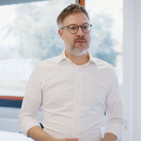 Ein Mann in einem weissen Hemd steht an einem hellen Fenster, die Hände in Taillenhöhe und die Ärmel bis zu den Ellbogen aufgekrempelt.