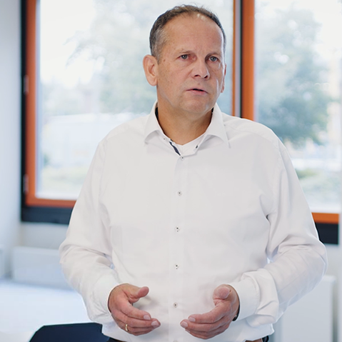 Ein Mann mittleren Alters in weißem Hemd gestikuliert mit beiden Händen in einem hellen Büro vor großen Fenstern.