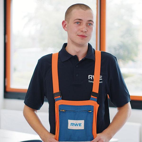 Ein Arbeiter steht drinnen in dunkelblauem Polo und orangefarbenen Hosenträgern mit blauer RWE-Tasche; im Hintergrund Fenster mit orange Rahmen.