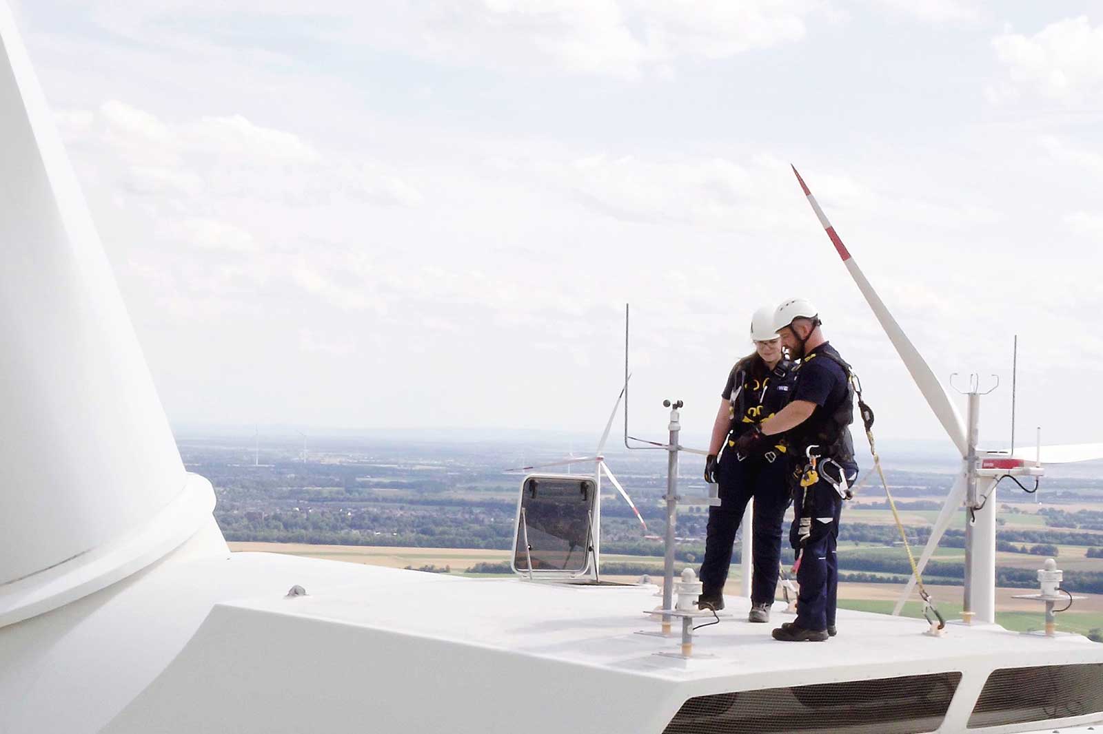 Zwei Personen stehen auf der Spitze eines Windrades und genießen den Blick auf die Landschaft mit Feldern und Wolken.