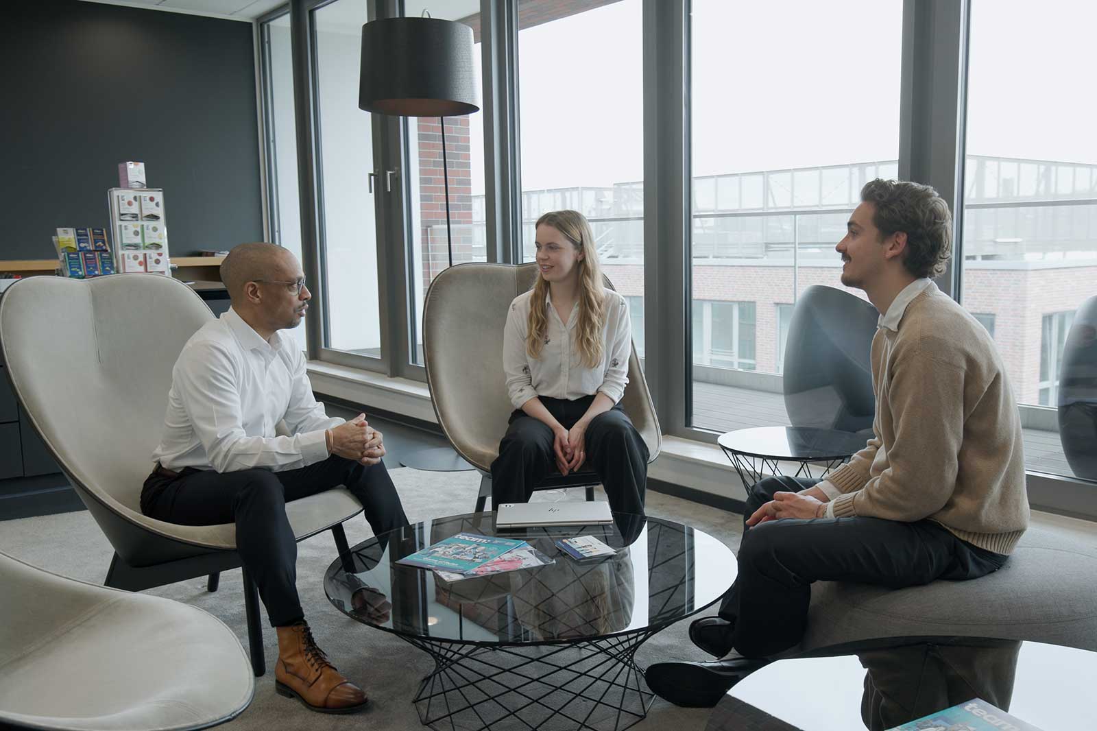 Drei Personen sitzen in modernen Stühlen in einem Büro. Ein runder Glastisch steht vor ihnen, umgeben von großen Fenstern.