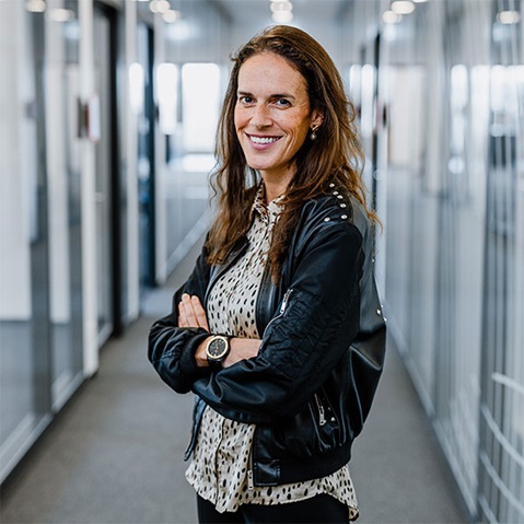 A person stands in a modern office corridor, wearing a black jacket and a spotted shirt. Portrait of Giordana Doppstadt.