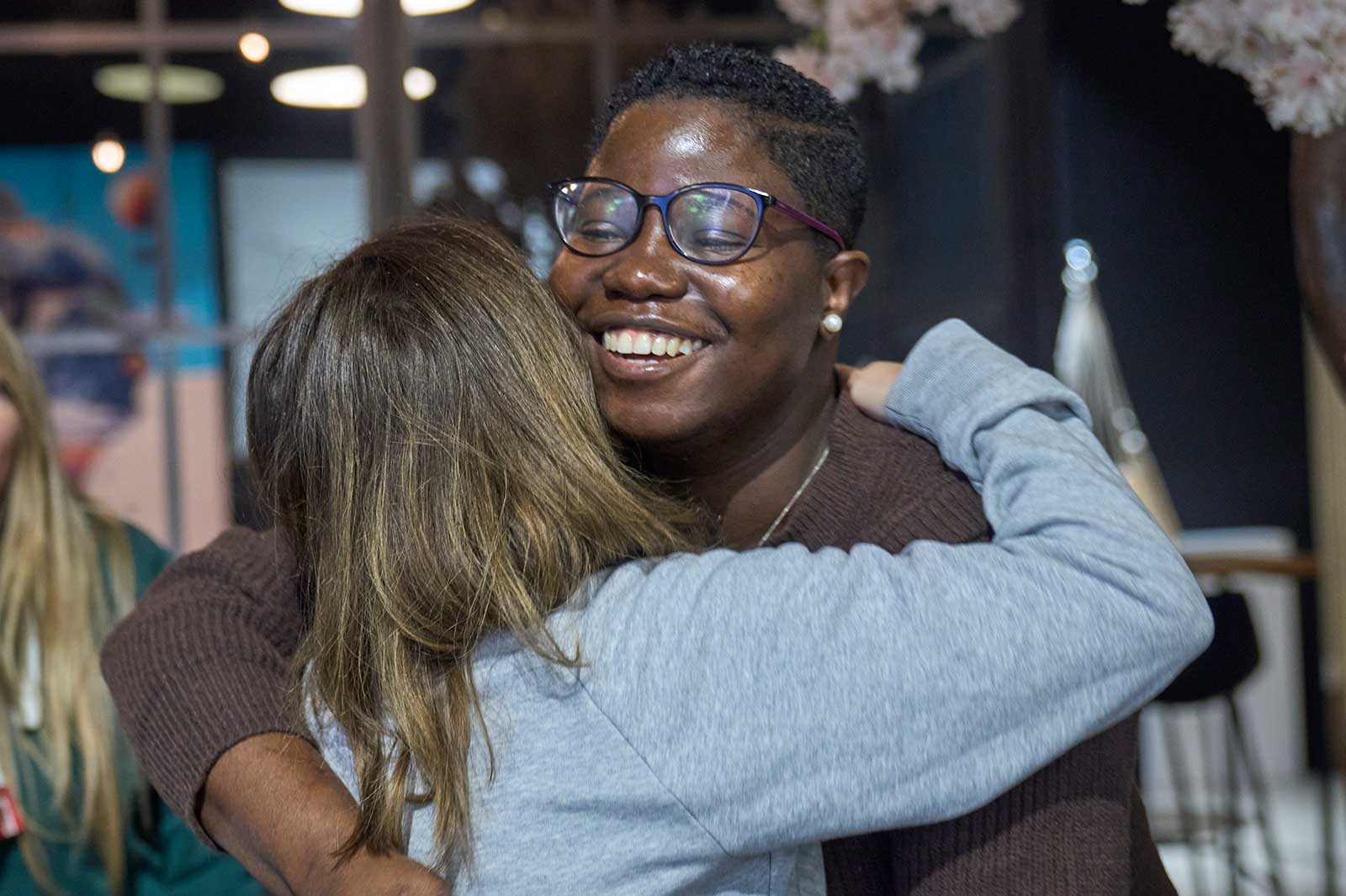Eine Frau umarmt einen Mann in einer lässigen Umgebung, umgeben von verschwommenen Hintergründen und sanftem Licht.