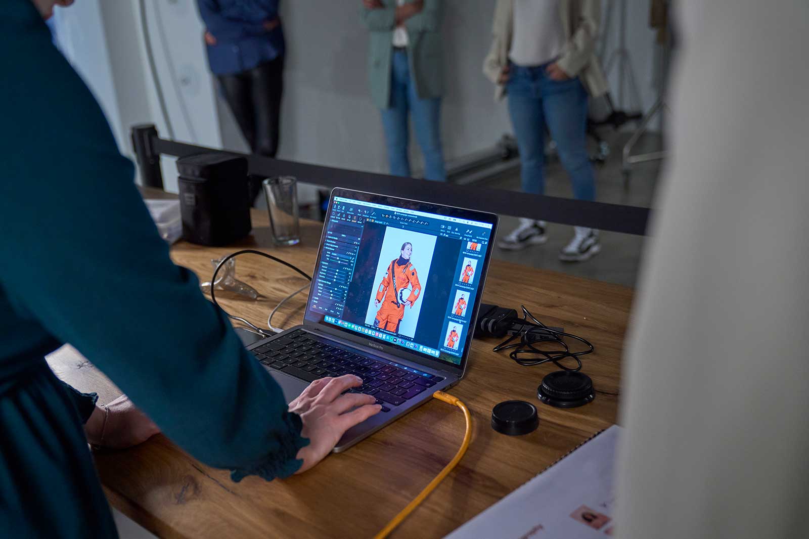 A person edits photos on a laptop placed on a wooden table. People are visible in the background.