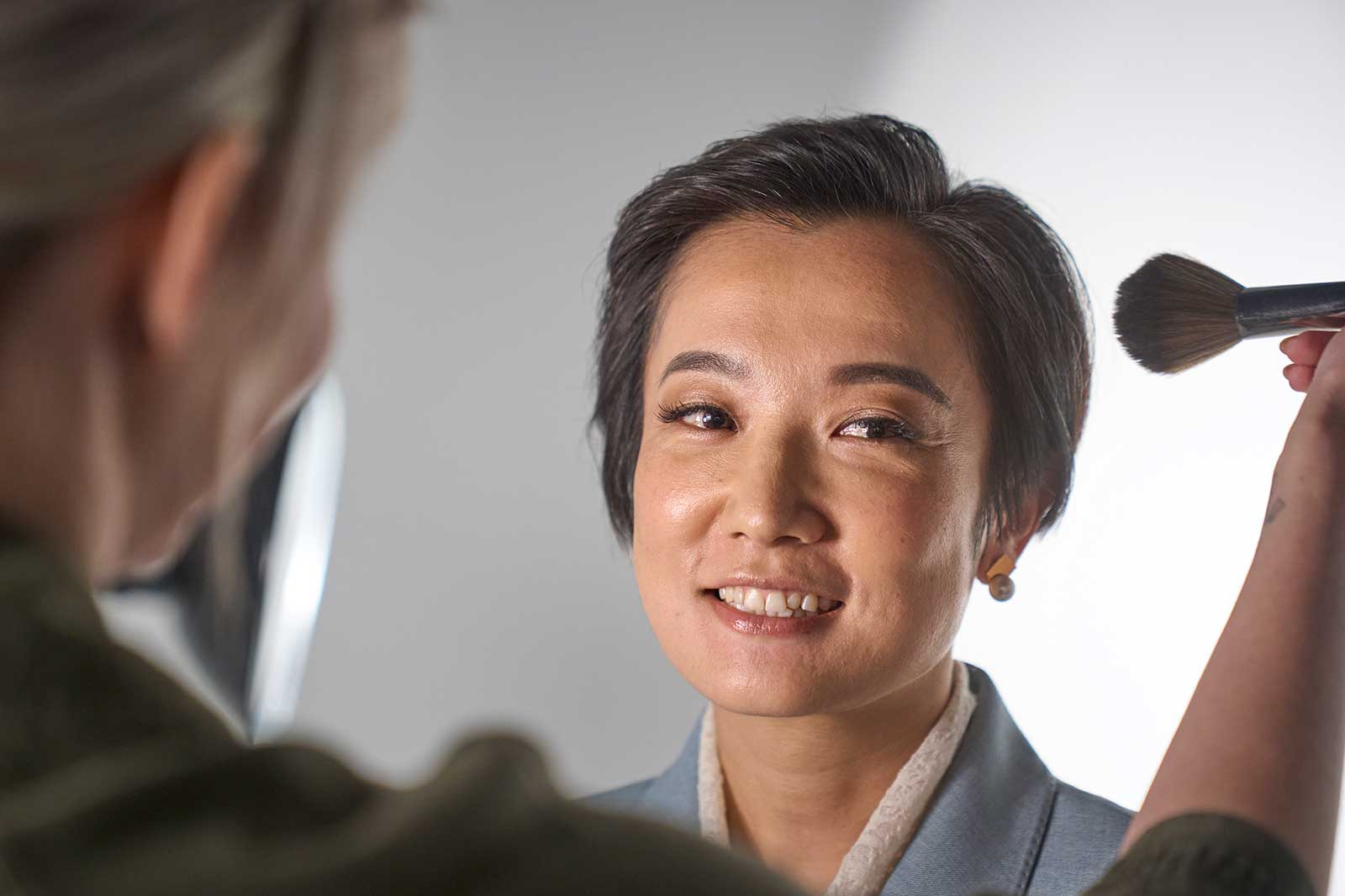 Eine Person mit kurzer Frisur wird mit einem Pinsel geschminkt. Der Hintergrund ist hell und neutral.
