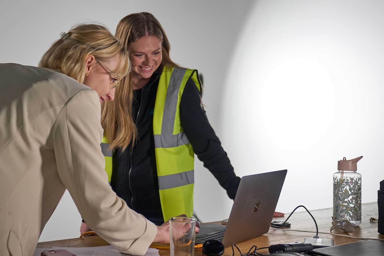 Zwei Frauen besprechen sich über einem Schreibtisch mit einem Laptop, eine trägt eine Warnjacke und eine Wasserflasche ist in der Nähe.