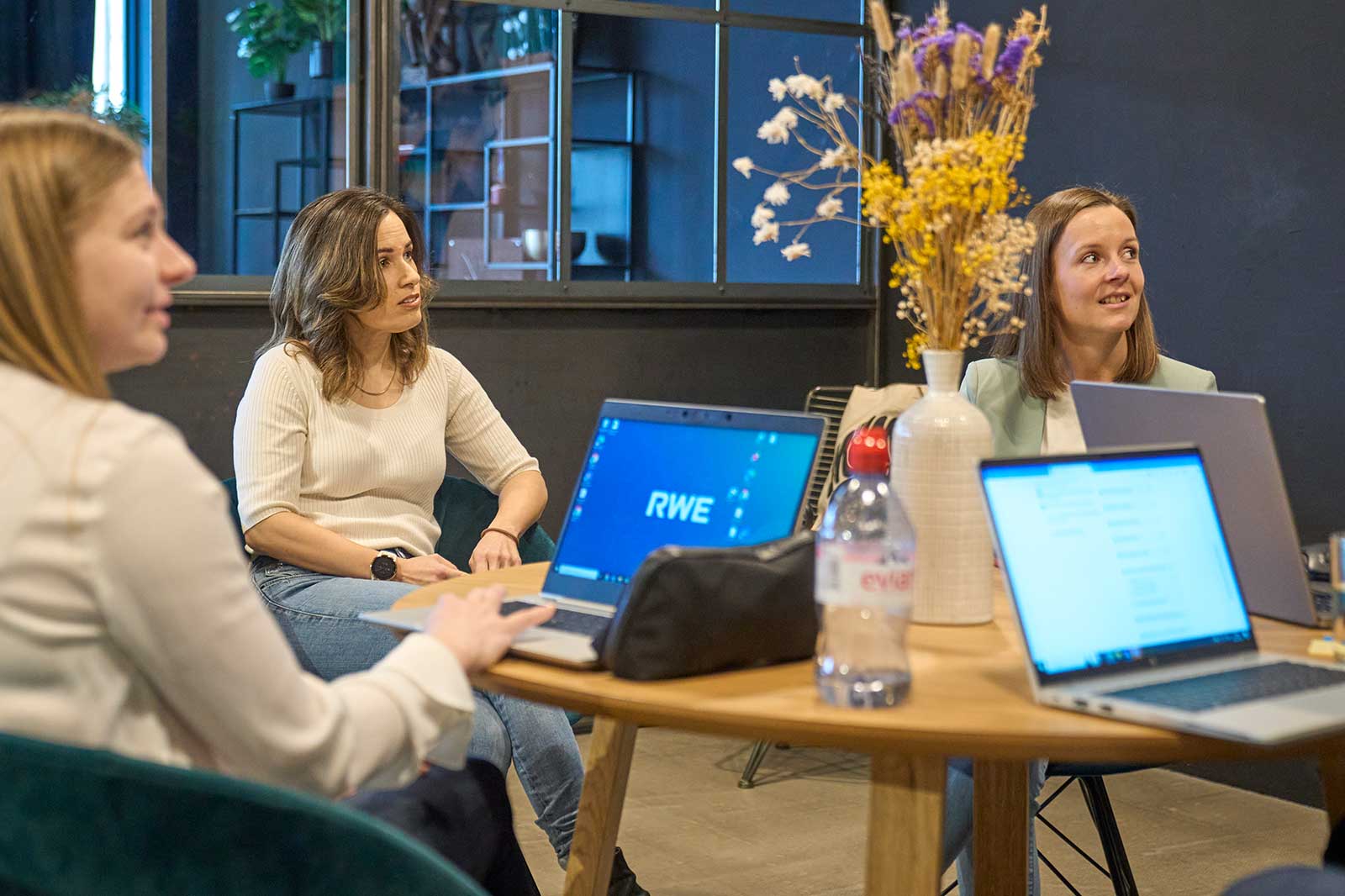 Three people sit at a table surrounded by laptops and a vase of dried flowers.