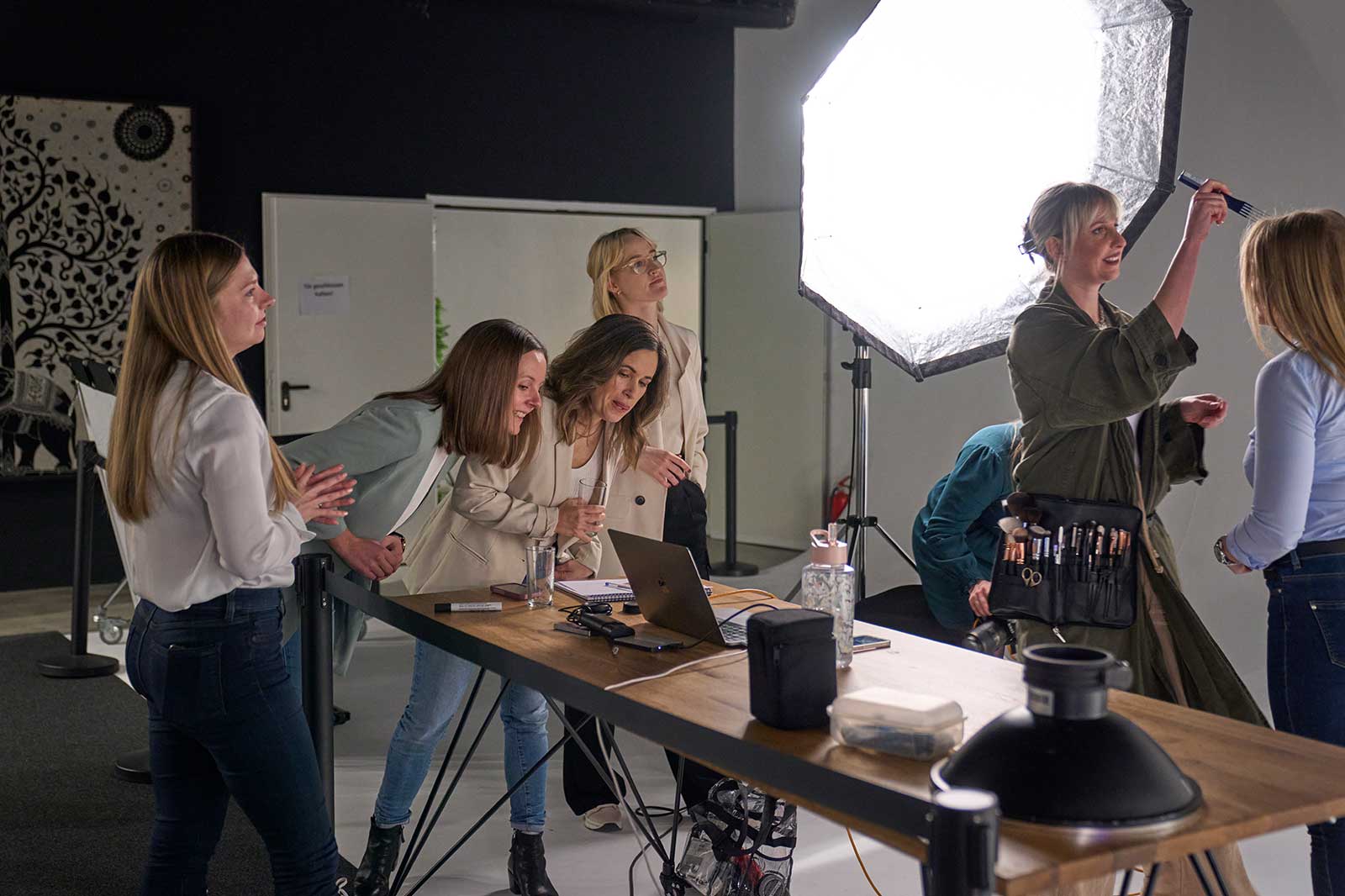 A group of women stands by a table with makeup and equipment. A large light illuminates the scene.