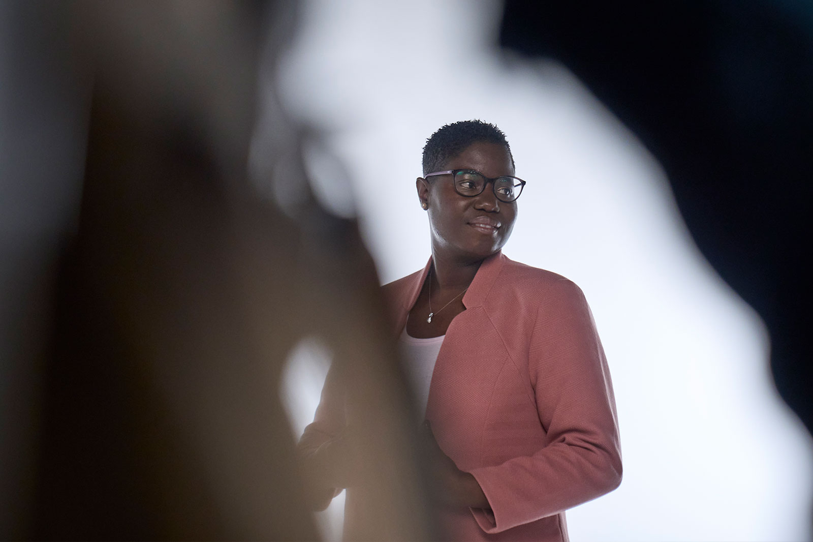 A person stands in the foreground against a light background, wearing a pink blazer.