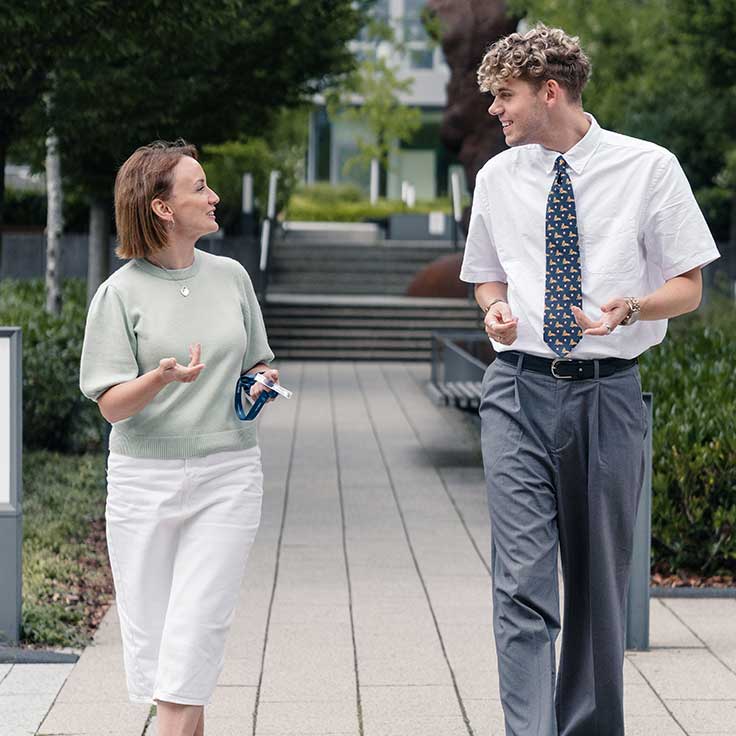 Eine junge Frau in einem hellgrünen Pullover und weißen Hosen unterhält sich mit einem Mann in einem weißen Hemd und gemusterten Krawatte im Freien.