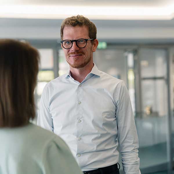 Ein Mann trägt ein hellblaues Hemd und steht einer Frau gegenüber, während er sich in einer modernen Büroumgebung unterhält. Portrait von Jacob Meins.