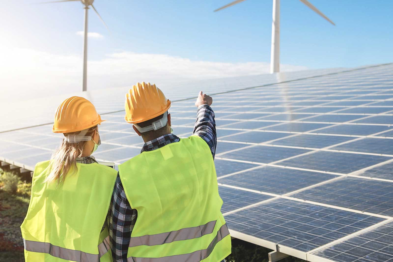 Zwei Ingenieure in gelben Westen und Helmen beobachten Solarmodule und besprechen die nahegelegene Windturbine unter einem hellen Himmel.