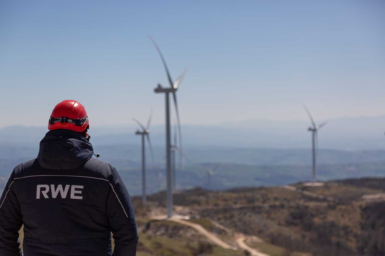 Ein Arbeiter mit einem roten Helm steht und blickt auf mehrere Windkraftanlagen an einem sonnigen Tag vor blauem Himmel.
