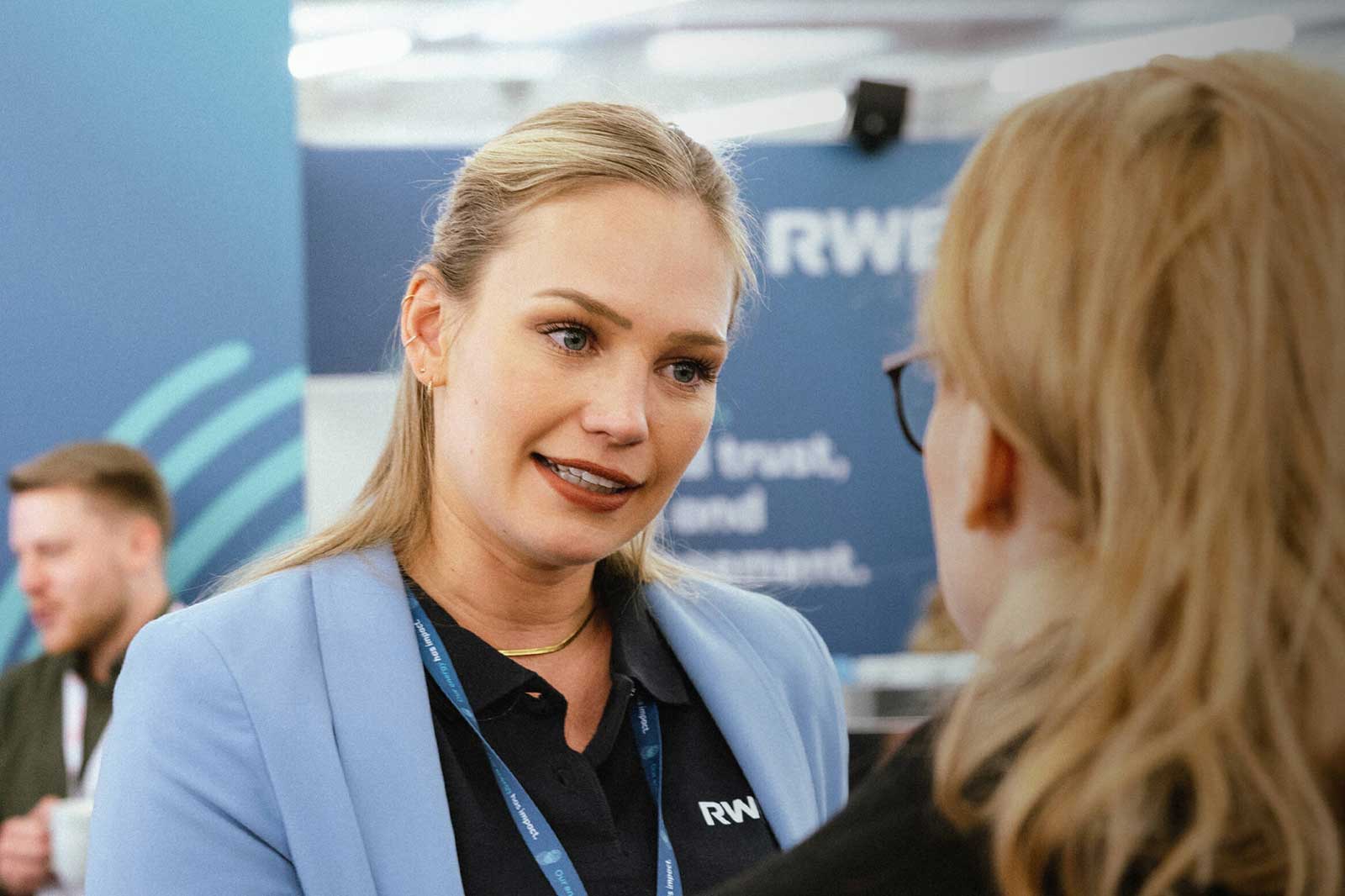 Eine Frau in einem blauen Blazer spricht mit einer anderen Person bei einer Veranstaltung, im Hintergrund sind Markenmaterialien sichtbar.