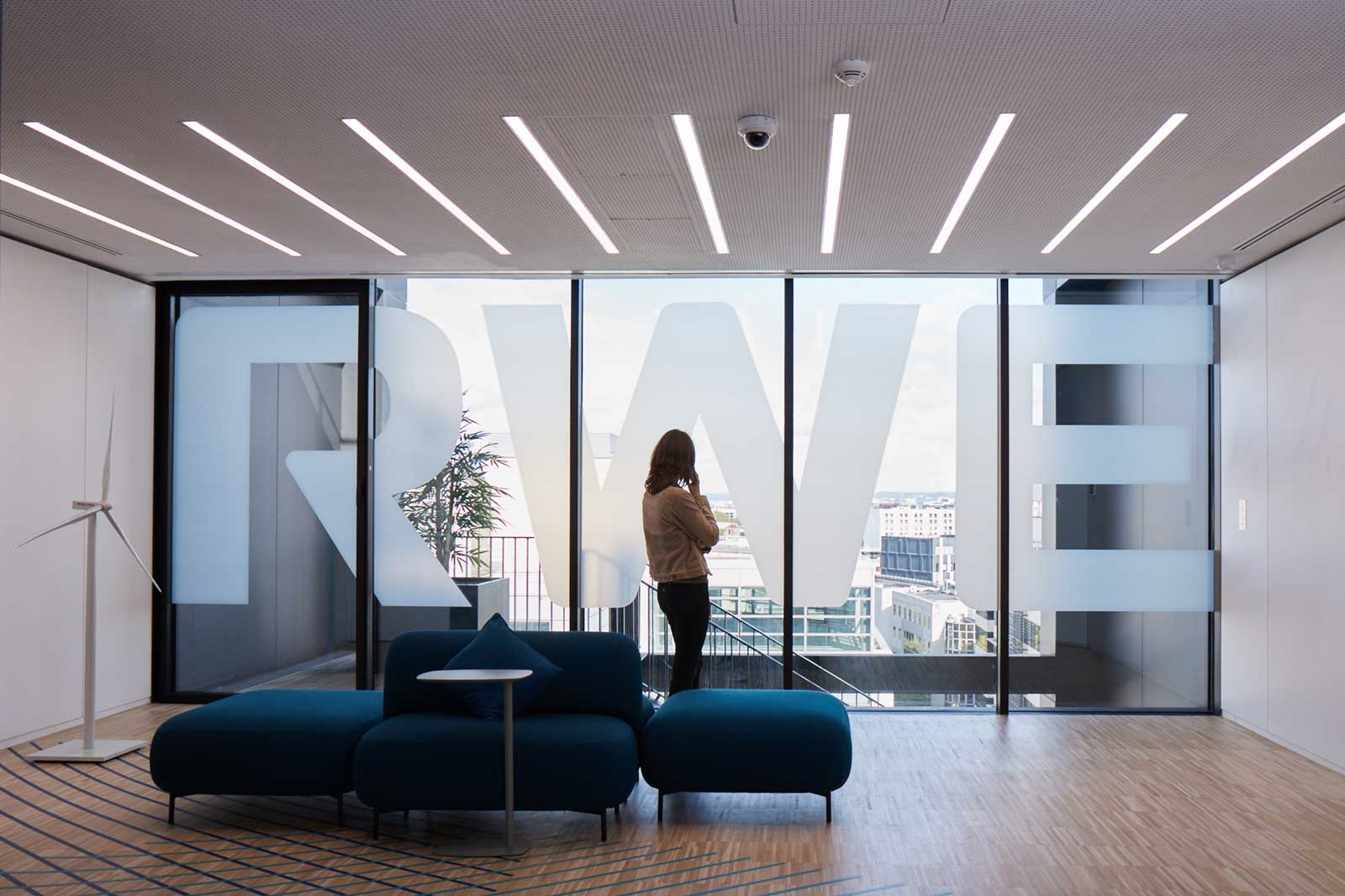 A woman gazes out of a large glass window with the letters 'RWE' etched on it in a modern office space featuring a dark blue sofa.