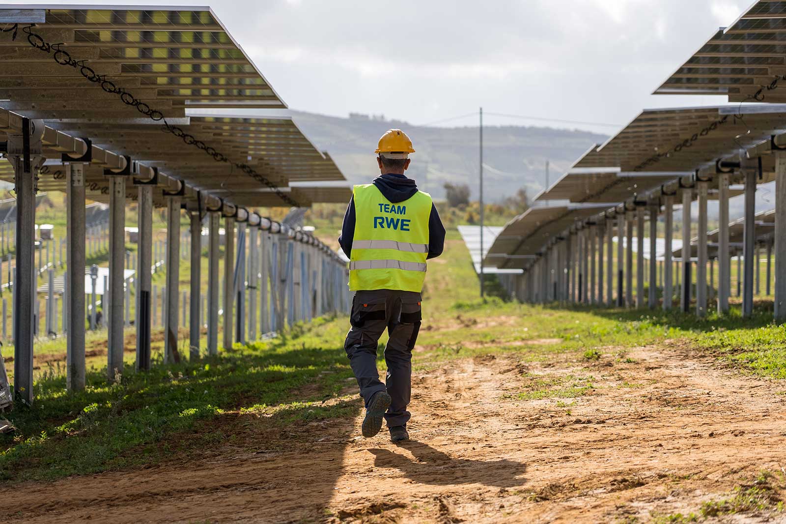 A worker in a bright vest walks between rows of solar panels on a sunny, green landscape.