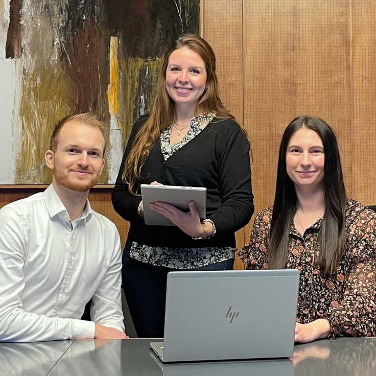 Drei Personen in einem Besprechungszimmer, mit einem Laptop und einem Notizblock, vor einem abstrakten Gemälde.