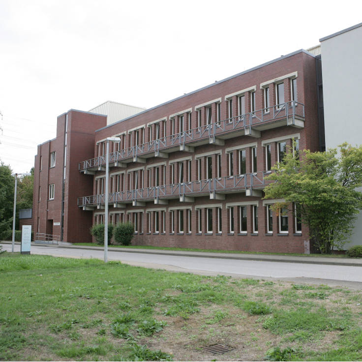 Ein modernes Backsteingebäude mit mehreren Balkonfenstern und Grünflächen davor, gelegen an einem Gehweg.