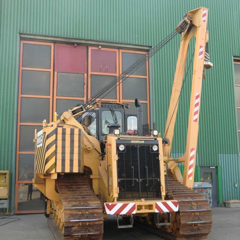 Ein großer gelb-schwarzer Raupenbulldozer mit einem Kranarm, vor einer Fabrik mit grünen Wänden positioniert.