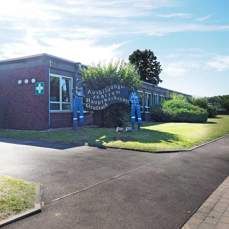 Ein Gebäude mit dem Namen Ausbildungszentrum Hauptwerkstatt Gelefrath und cartoonhaften Arbeiterfiguren davor in einer sonnigen Umgebung.