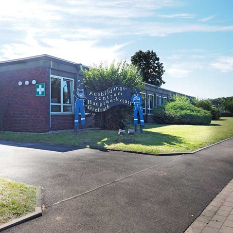 Ein Gebäude mit dem Namen Ausbildungszentrum Hauptwerkstatt Gelefrath und cartoonhaften Arbeiterfiguren davor in einer sonnigen Umgebung.