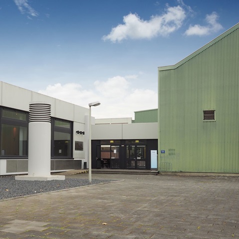 Ein modernes Gebäude mit einer grünen Fassade und einem Glaseingang, gelegen in einem gepflasterten Bereich unter einem bewölkten Himmel.