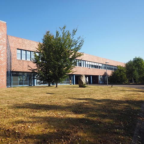 Ein modernes Backsteingebäude umgeben von Grünflächen, mit großen Glasfenstern unter einem klaren blauen Himmel.