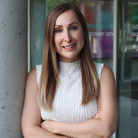 A person with long, straight hair stands by a column. They are wearing a sleeveless, textured top and have their arms crossed.