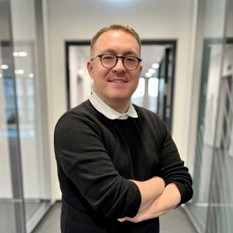 A man in a black sweater and white collar stands with crossed arms in a modern office.