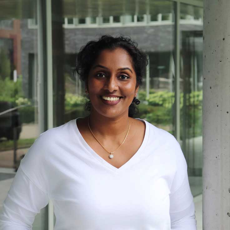 A person in a white top stands in front of a modern building with large glass windows and landscaped greenery.