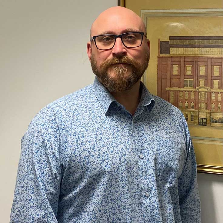 Eine Person in einem blauen, floralen Hemd steht vor einem gerahmten Bild eines Gebäudes.