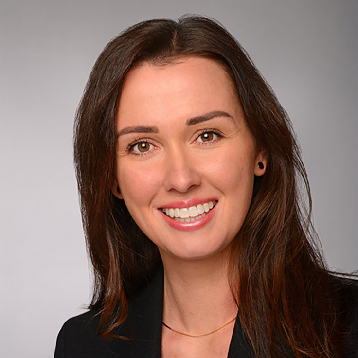 A woman with long brown hair is wearing a black jacket and a gold necklace. The background is light grey.
